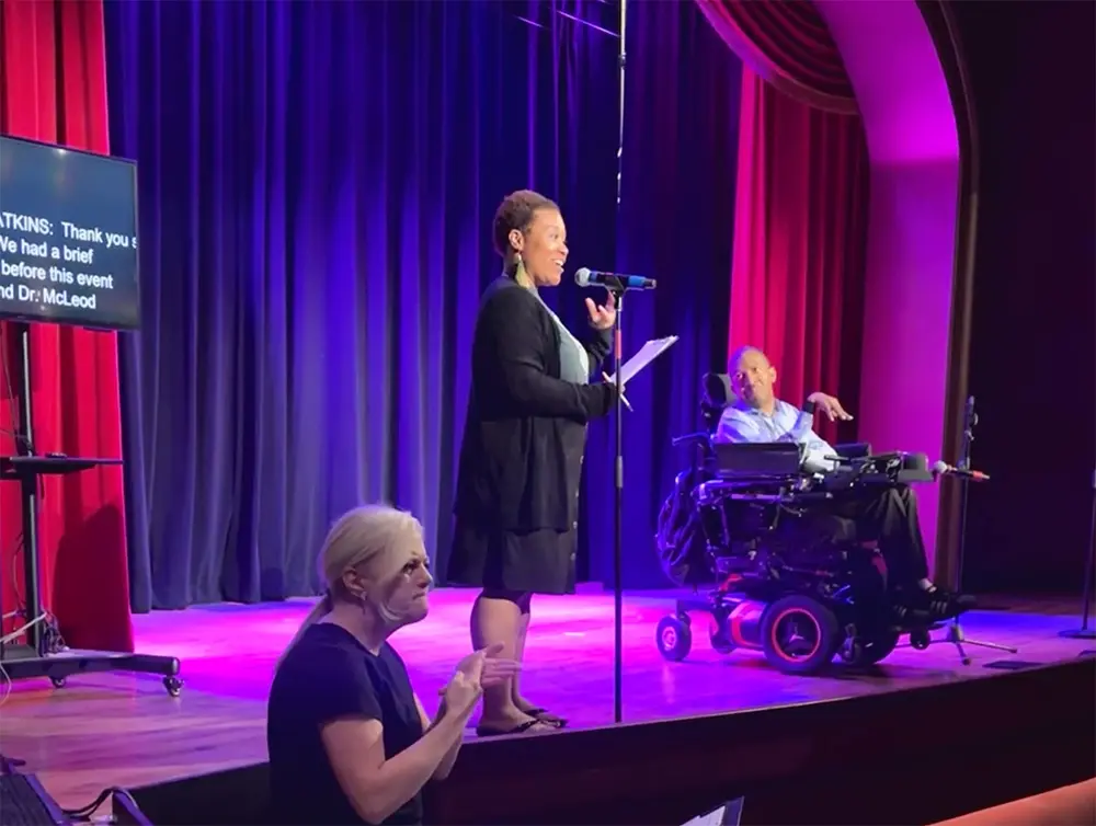 A Black woman standing on stage, speaking into a microphone. Beside her is a Black man in a powerchair. On the bottom left is a white woman with her hair pulled back. She is an ASL interpreter. Live captions can be seen behind them.
