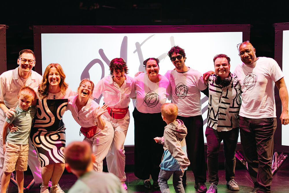 Eight adults standing together, smiling. One child has joined them for the photo. 2 children are facing them and standing.