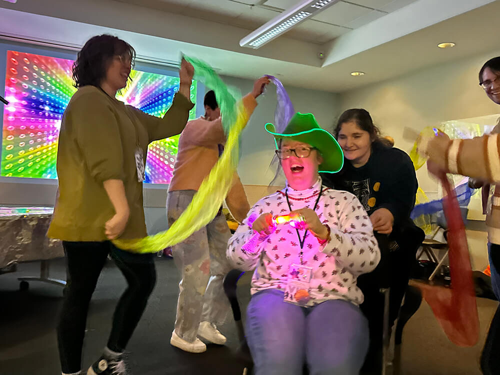 A person in a wheelchair, wearing a long sleeve shirt, jeans, and a green cowboy hat. They are surrounded by four people holding scarves, presumably dancing with them around said person.