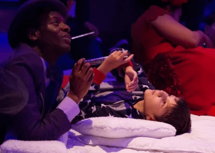 A Black man in a suit and hat, laying with a young boy in his pajamas. It appears this is during a live performance of sorts.