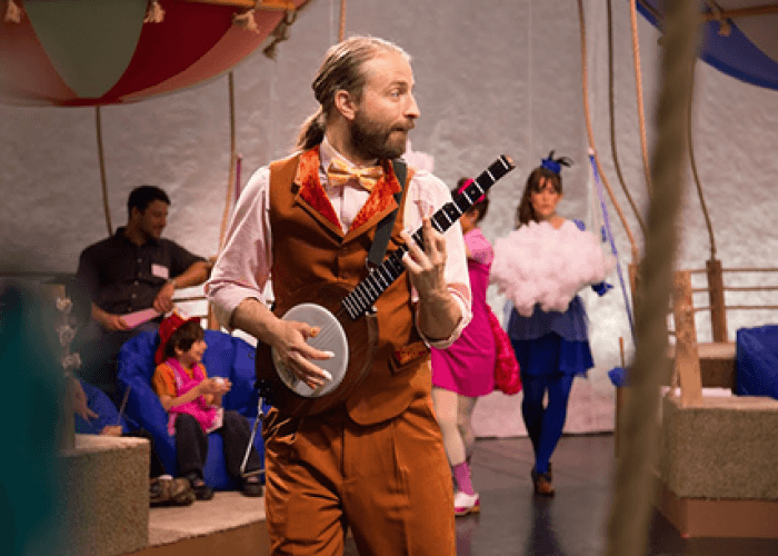 A man in costume, playing a banjo. Three people are in the background.