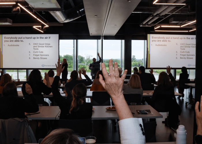 Audience members raise their hand, being engaged with a public speaker. They are in a conference room setting.