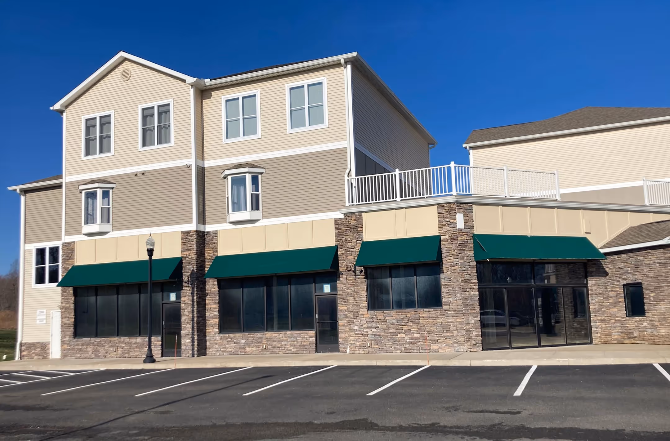 Two-story building with a stone facade and green awnings, featuring a storefront. The upper floor has beige siding and white-framed windows.