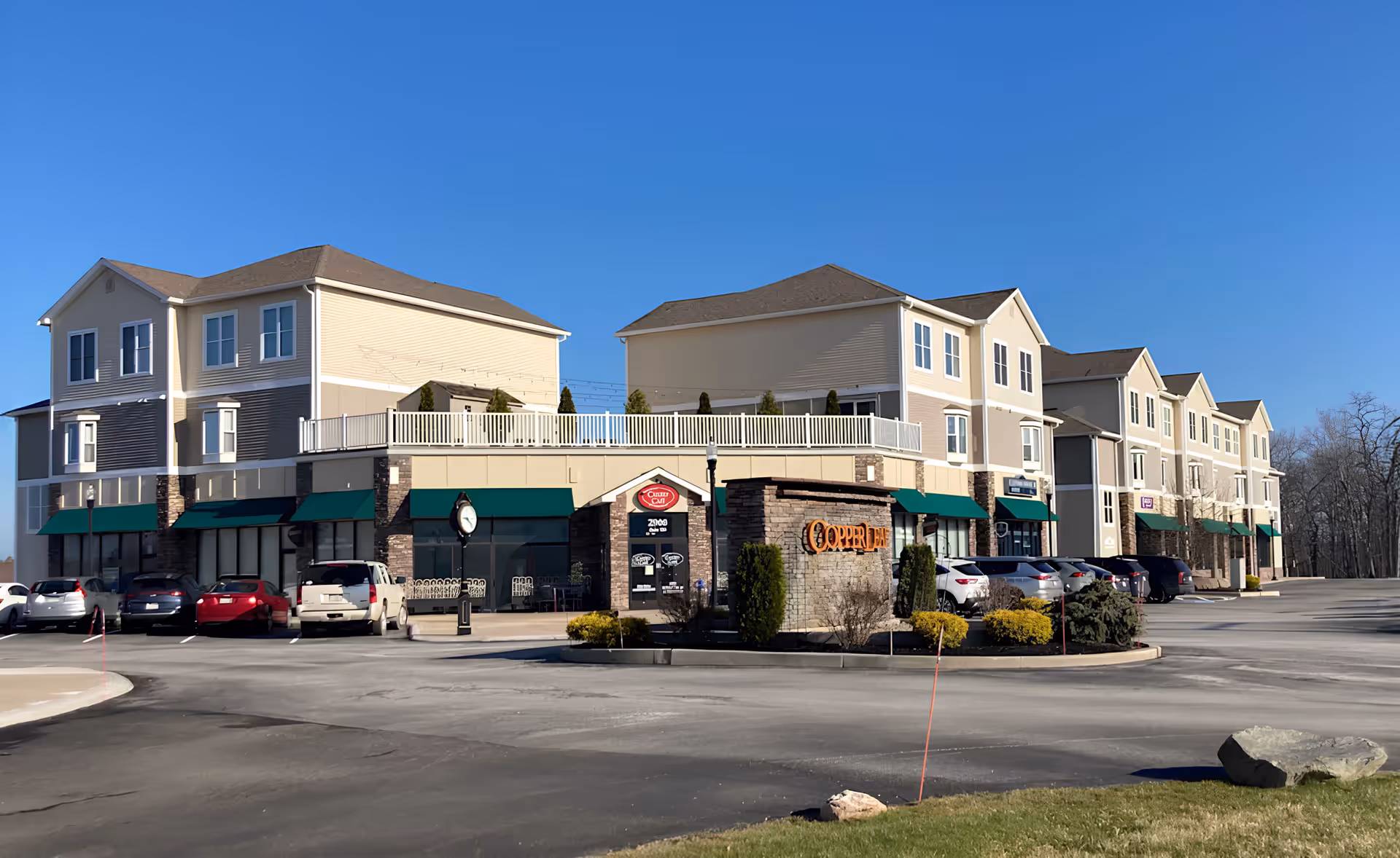 Three-story beige commercial building under a clear blue sky. The ground floor has retail shops with green awnings; parked cars are in front. Tone is calm and sunny.