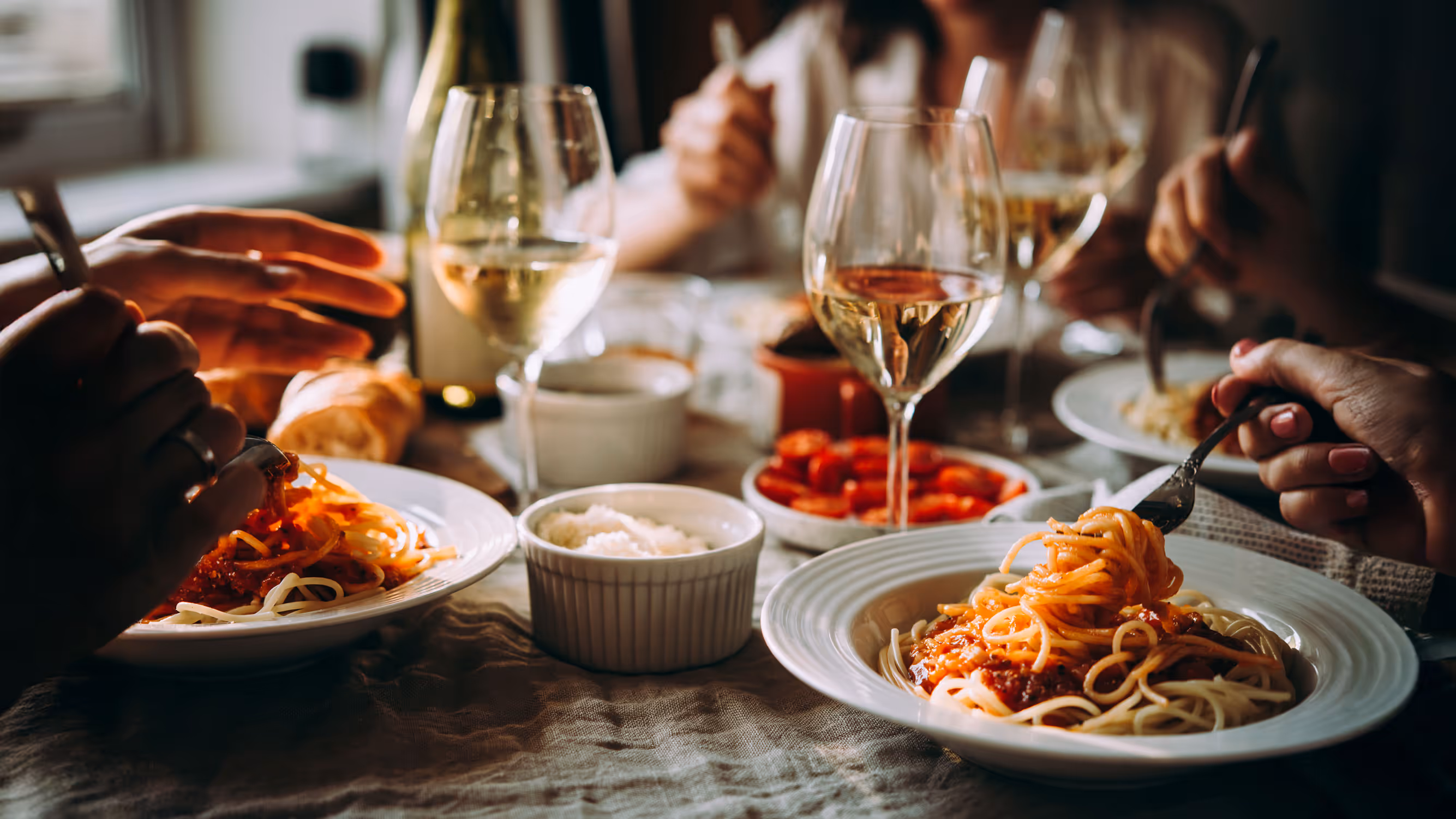 A cozy dinner scene with several people enjoying spaghetti and wine. Plates of pasta, glasses of white wine, and bowls of grated cheese adorn the table.