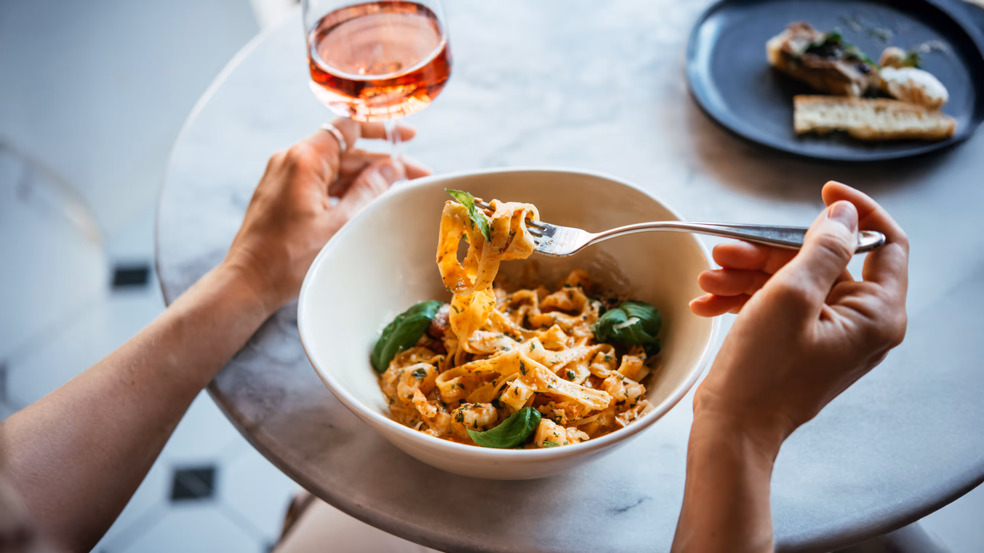 A person enjoys creamy pasta with basil in a white bowl, holding a forkful and a glass of rosé wine, creating a cozy dining atmosphere.