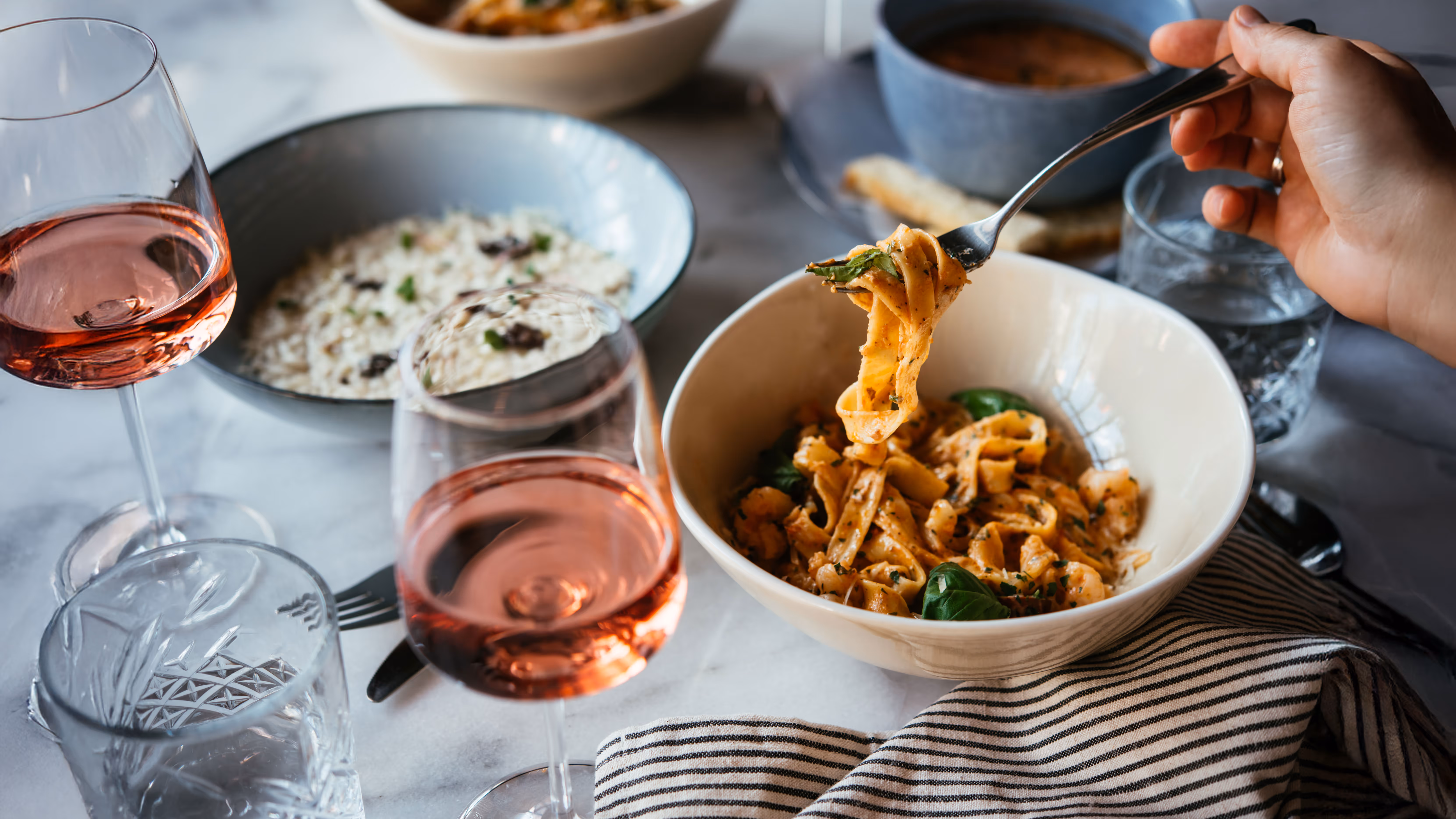 A cozy dining scene with a hand twirling pasta in a bowl. Nearby, rosé wine glasses and dishes create a warm, inviting atmosphere on a marble table.