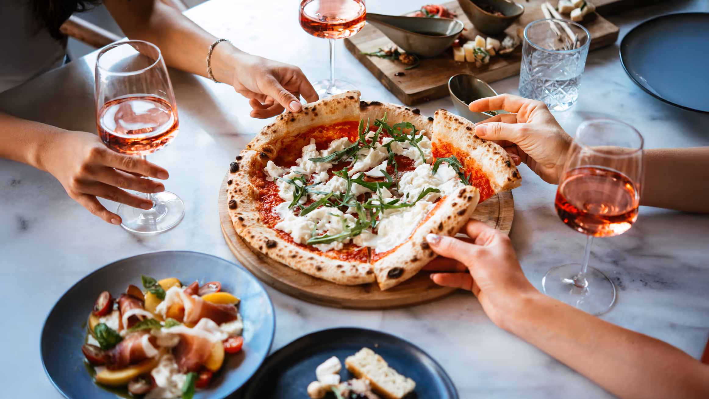 Aerial view of a shared pizza topped with arugula and cheese. Three hands reach for slices. Glasses of rosé and a colorful salad are nearby.