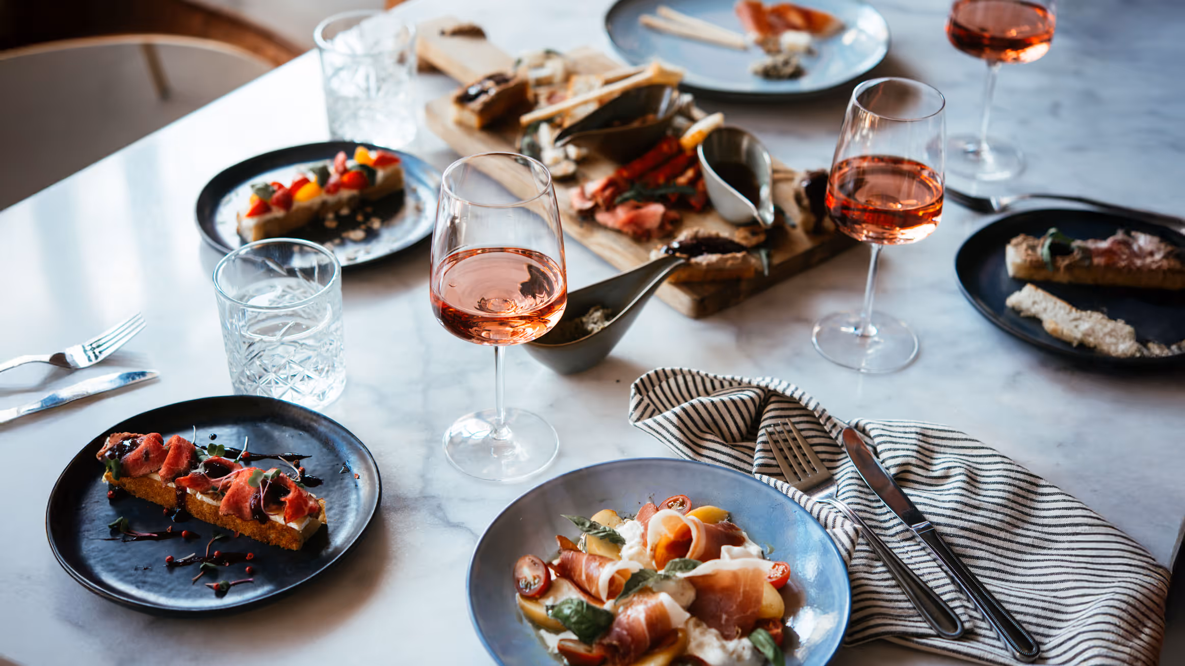 A stylish dining table with assorted gourmet appetizers on black plates, glasses of rosé wine, and a striped napkin, creating an elegant, inviting ambiance.