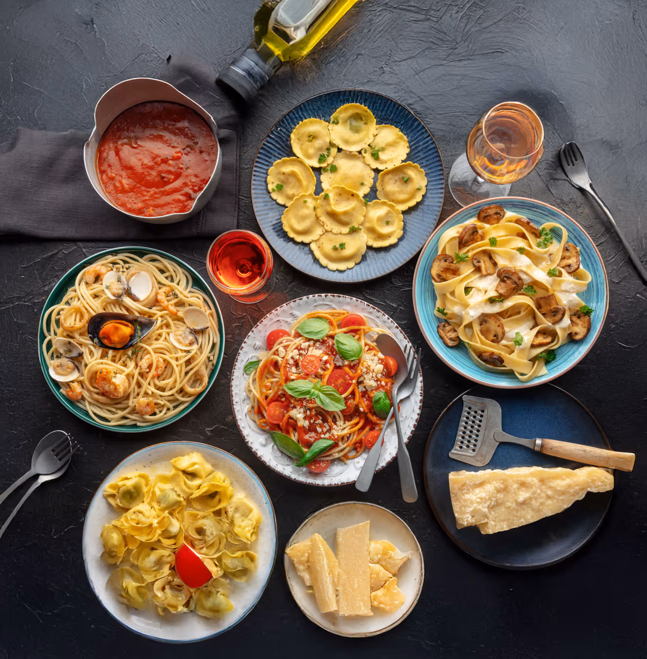A variety of pasta dishes on a dark surface, including spaghetti with clams, ravioli, tortellini, and pappardelle. Accompanied by Parmesan cheese, tomato sauce, and olive oil.