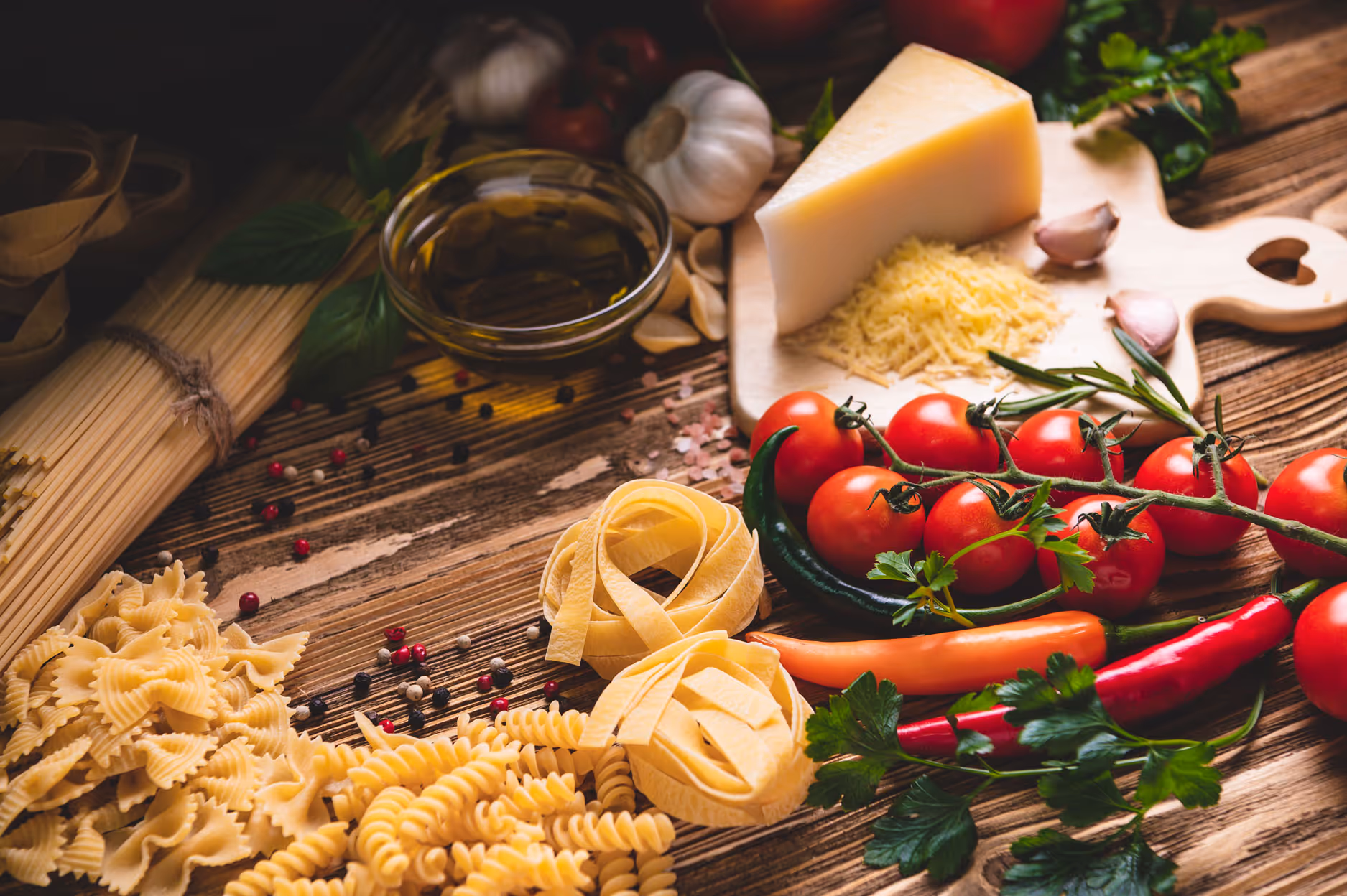 Assorted pasta, olive oil, tomatoes, spices, and cheese are arranged on a rustic wooden table, creating a warm and inviting Italian cooking scene.