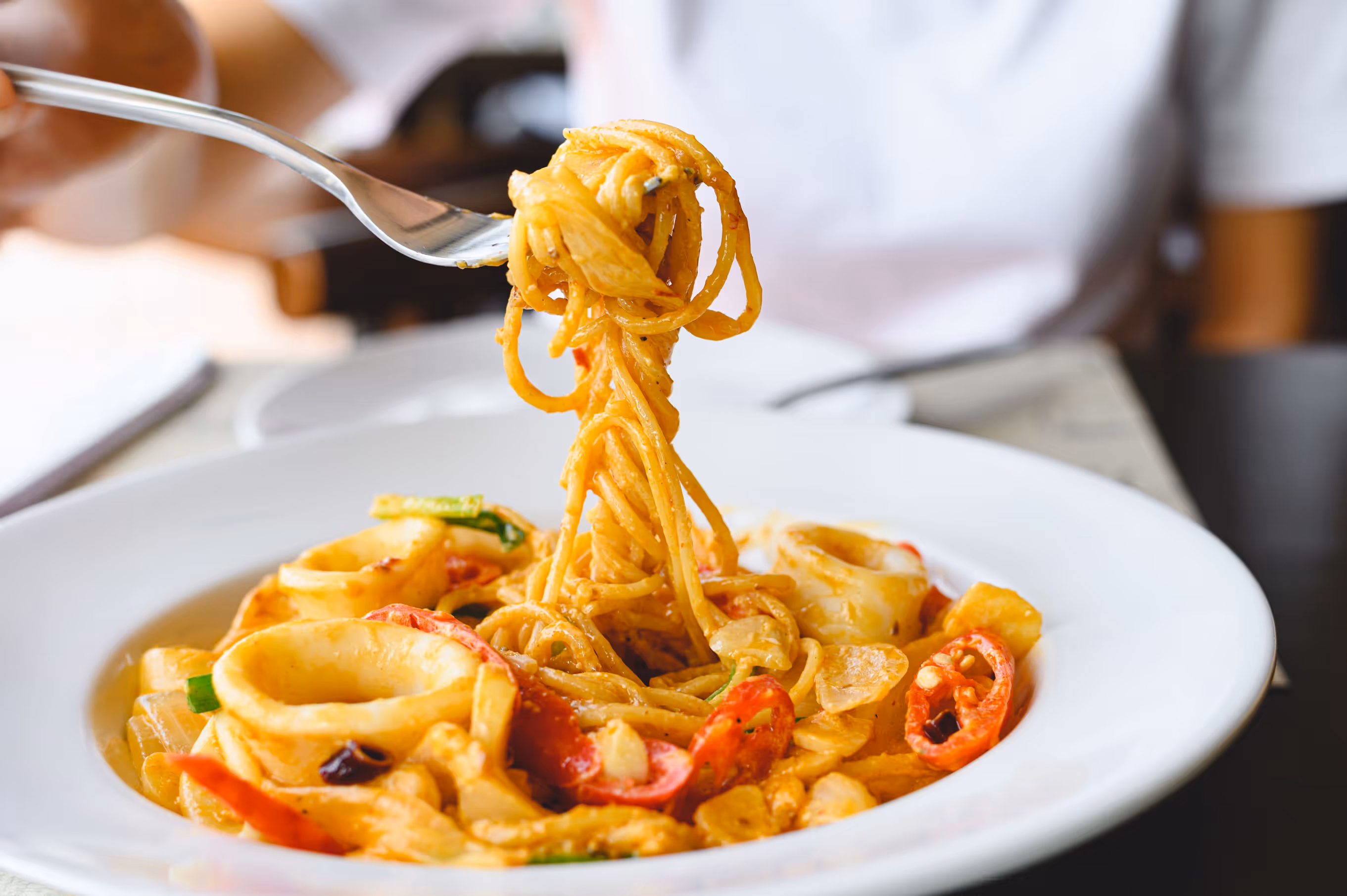 A vibrant seafood pasta dish with spaghetti, rings of calamari, and red pepper slices on a white plate. A hand twirls pasta with a fork, evoking warmth and appetizing scents.