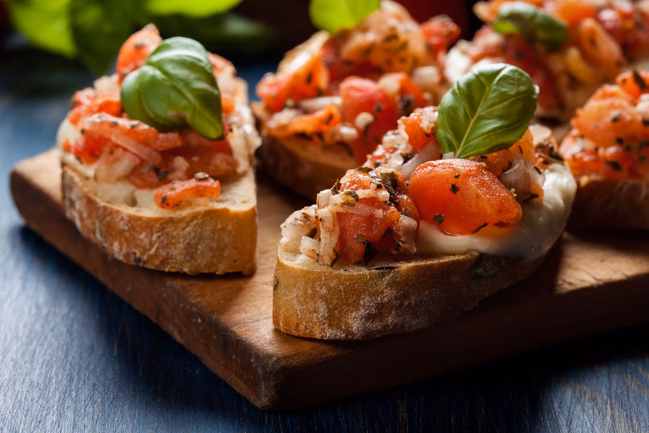Close-up of bruschetta topped with diced tomatoes, herbs, cheese, and basil leaves on crusty bread slices, arranged on a wooden board, conveying a fresh, appetizing feel.