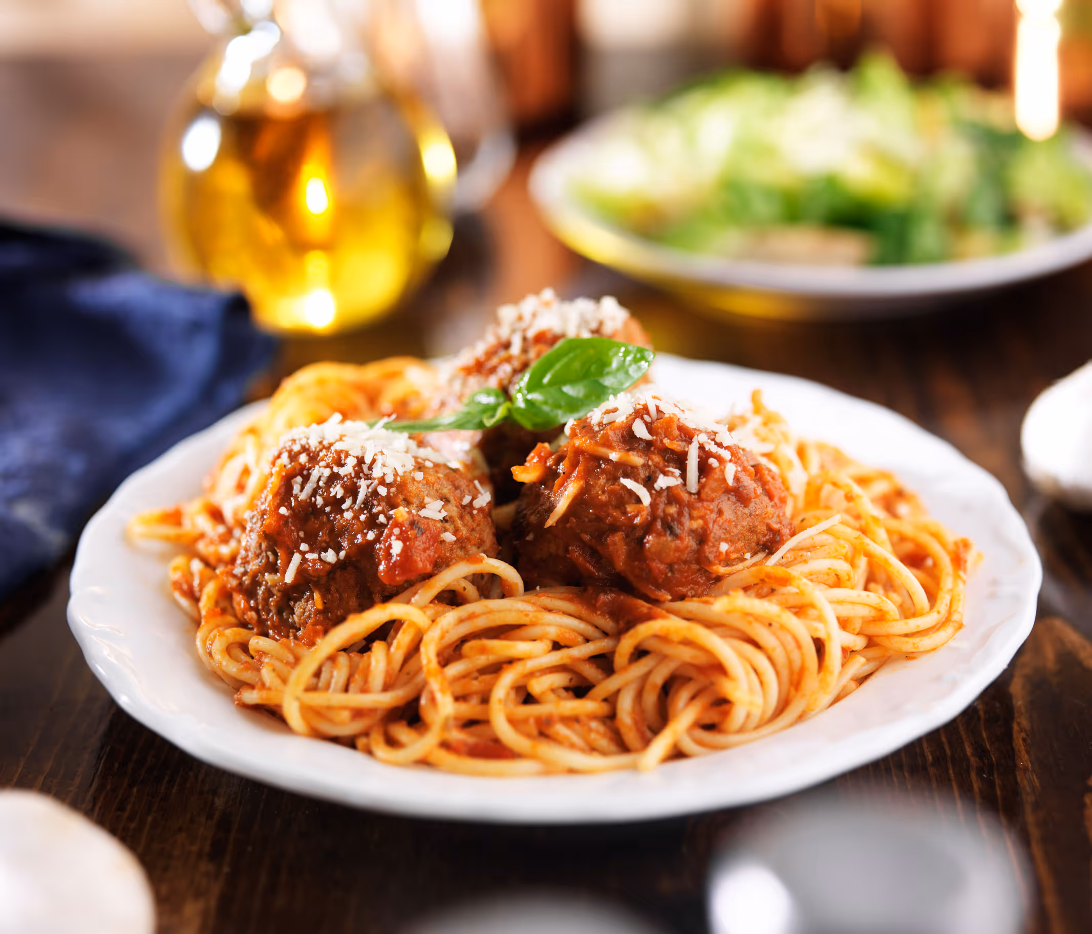 Plate of spaghetti with three meatballs, garnished with a basil leaf and cheese.