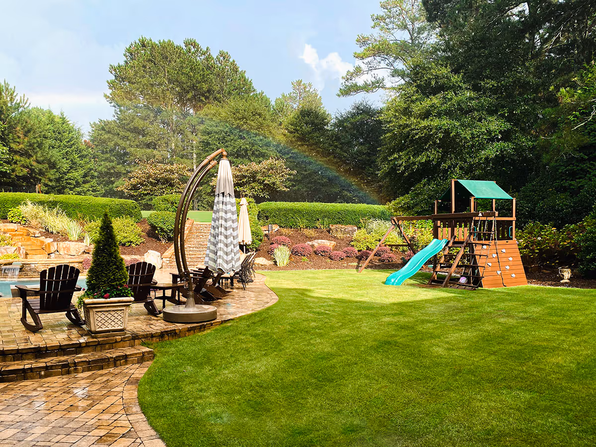 Backyard with green lawn, playground set with slide, patio seating area, and a visible rainbow over shrubs and trees.