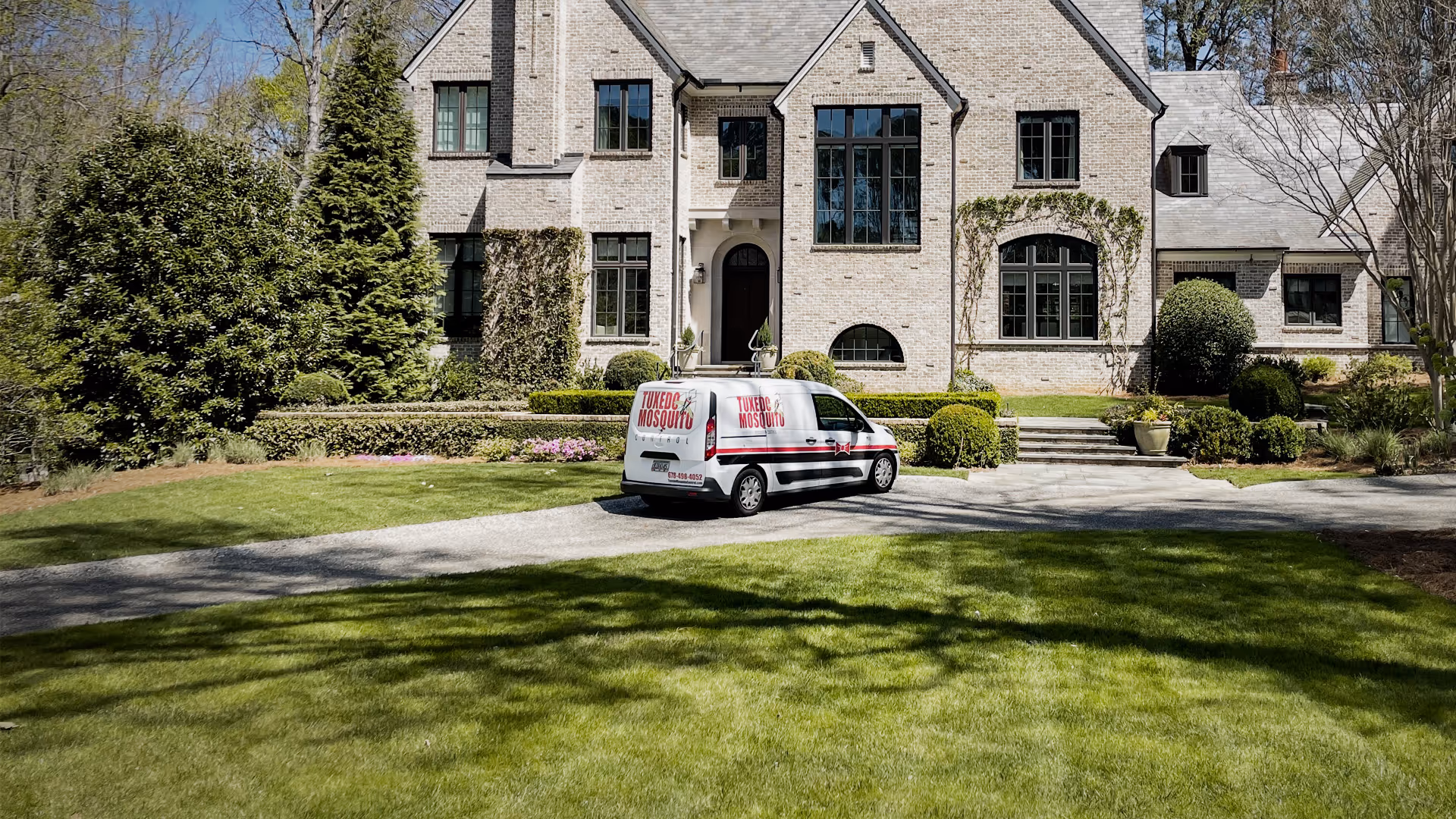 White Tuxedo Mosquito service van parked in front of a large brick house with landscaped yard.