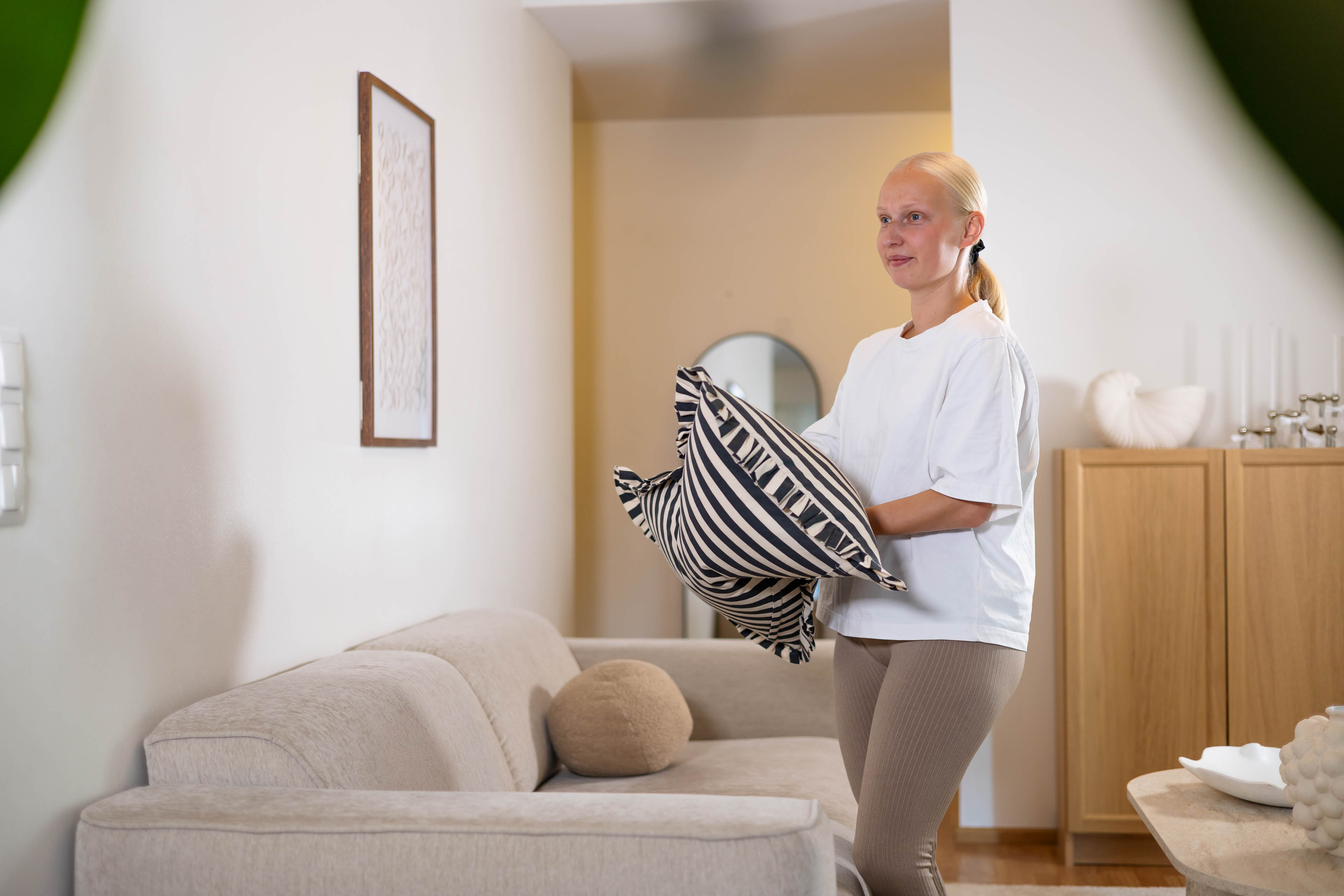 Nainen asettaa raidallisia tyynyjä vaaleanharmaalle sohvalle modernissa olohuoneessa.