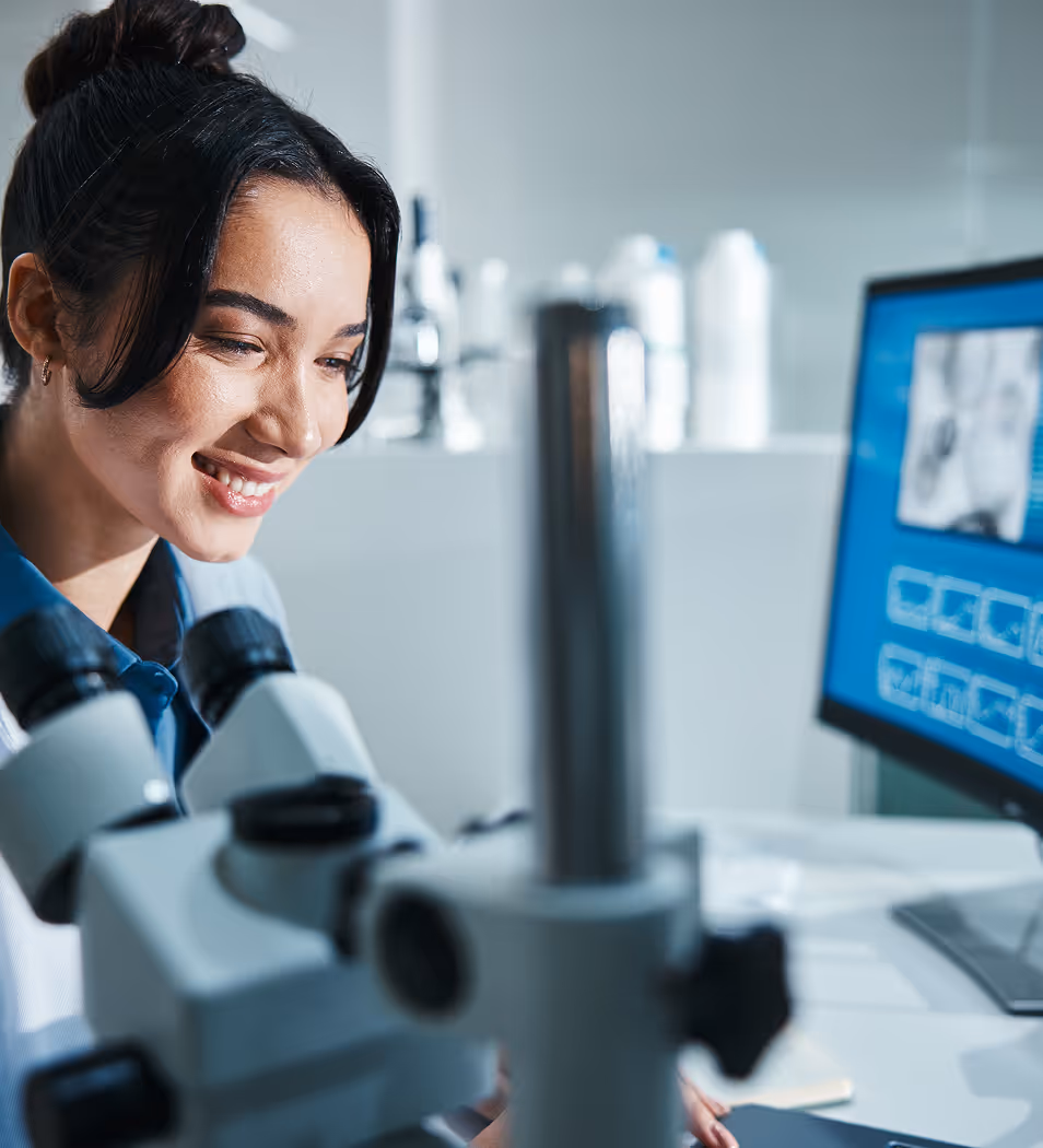 Female scientist smiling while working with a microscope in a laboratory with a computer screen displaying images in the background.
