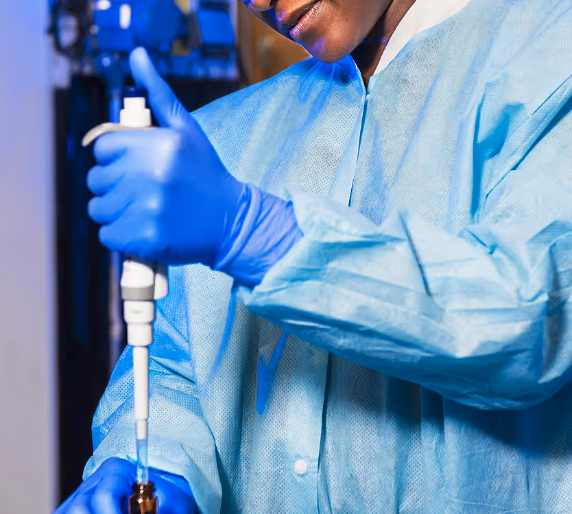 Scientist wearing blue gloves and a lab coat using a pipette to transfer liquid into a small brown bottle.