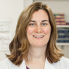 Smiling woman with shoulder-length brown hair wearing a white coat in an office setting.