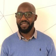 Man wearing black glasses and a blue sweater smiling against a white geometric wall background.