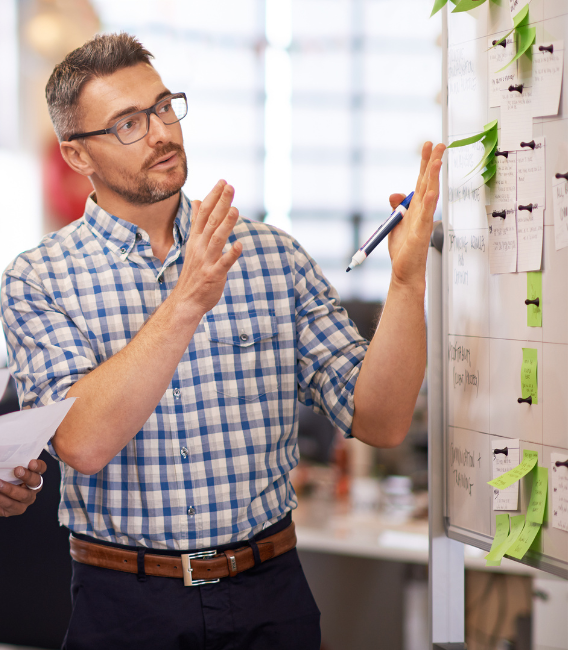 Man in a checkered shirt and glasses explaining or presenting ideas using a whiteboard with notes and sticky notes.