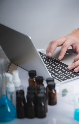 Person typing on a laptop keyboard with blurred bottles and laboratory equipment in the foreground.