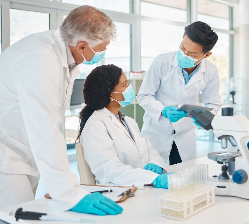 Three scientists wearing masks and lab coats discussing results in a bright laboratory with microscope and test tubes.