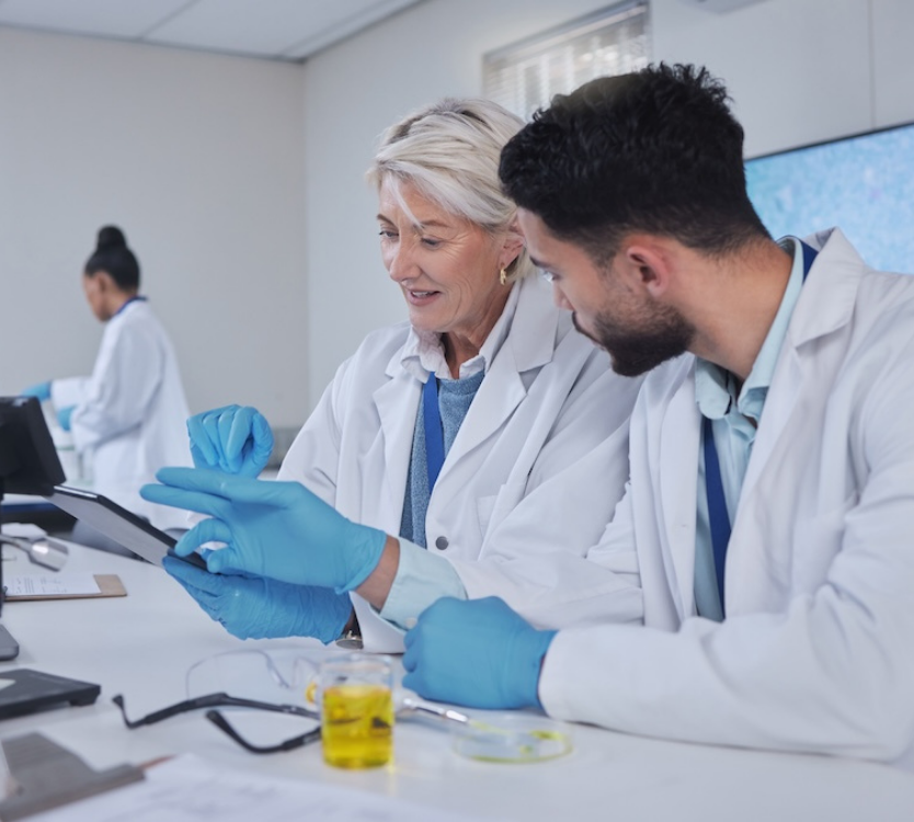 Two scientists in white lab coats and blue gloves examining a tablet together at a lab table with scientific equipment.