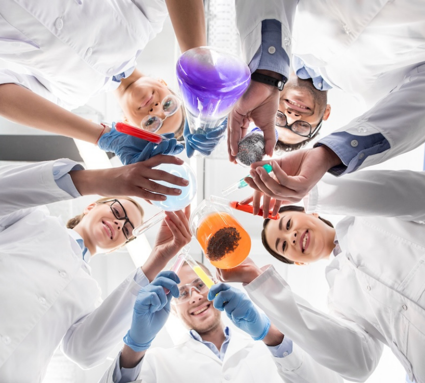 Group of smiling scientists in white lab coats holding colorful chemical liquids in glassware while looking down toward the camera.