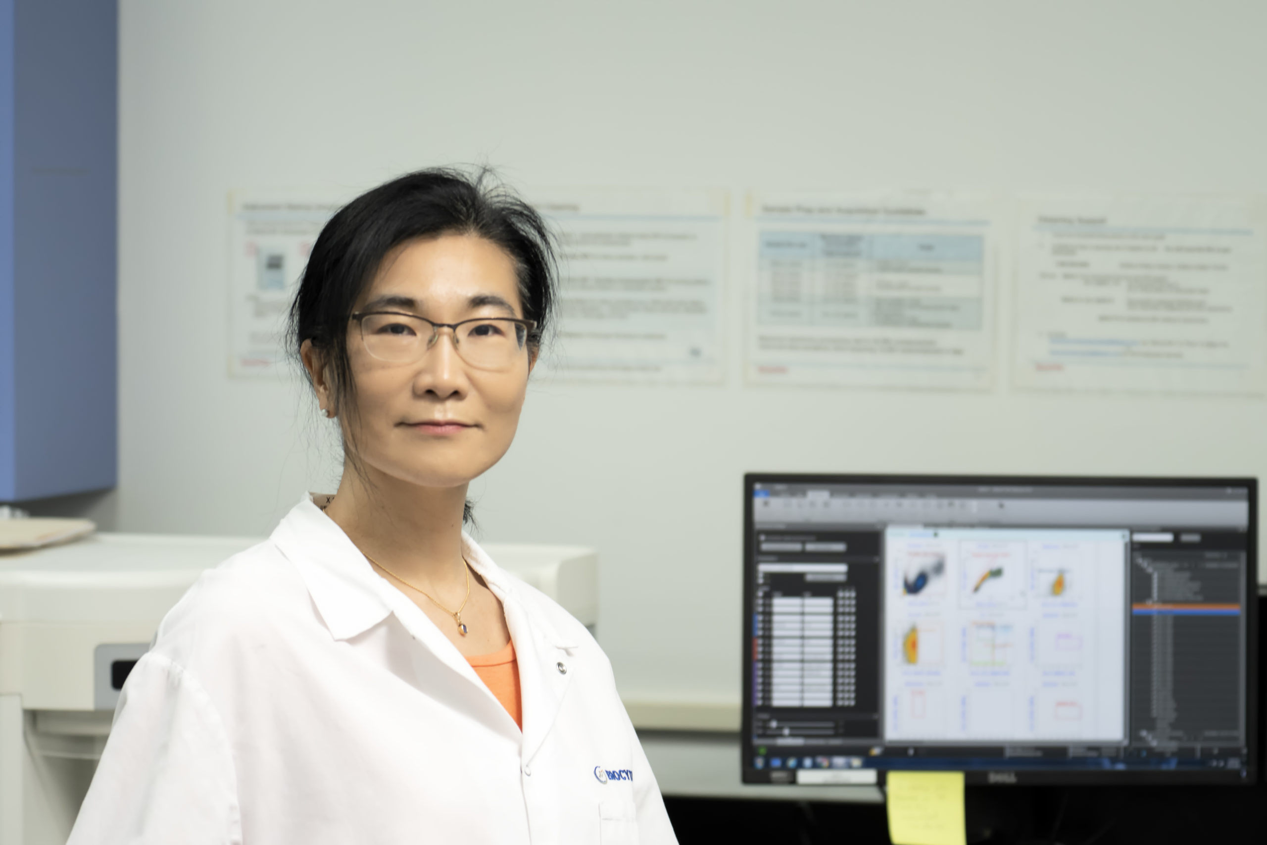 Female scientist wearing glasses and a white lab coat standing in a laboratory with a computer monitor displaying data in the background.