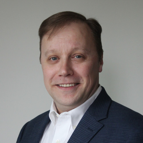Smiling man wearing a white shirt and dark blazer against a plain gray background.