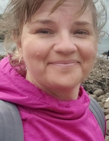 Close-up of a smiling woman wearing a pink hoodie and light gray backpack, outdoors with rocky terrain in the background.