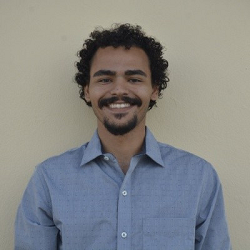 Smiling man with curly hair and a mustache wearing a light blue button-up shirt against a beige background.