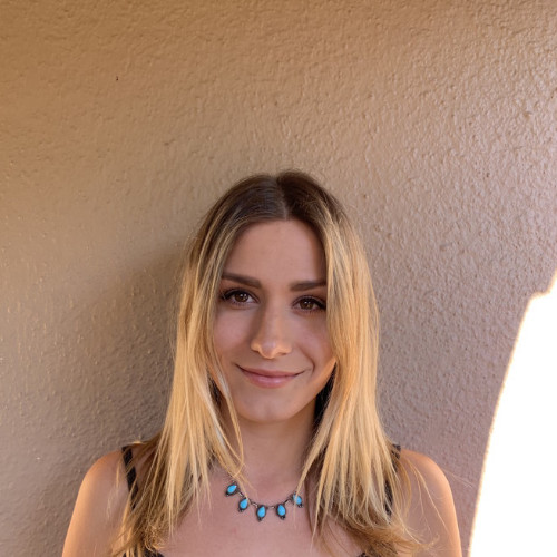 Portrait of a smiling young woman with long blonde hair wearing a turquoise necklace against a beige textured wall.