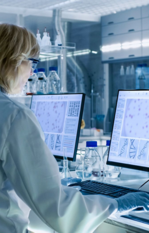 Scientist wearing glasses and gloves analyzing microscopic images on dual monitors in a laboratory.