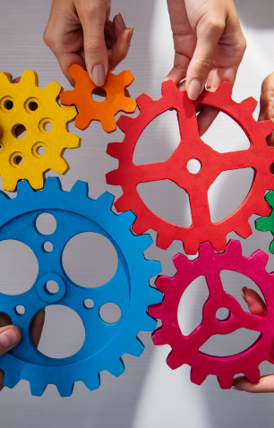Hands connecting colorful interlocking gears in orange, red, blue, pink, green, and yellow against a light background.