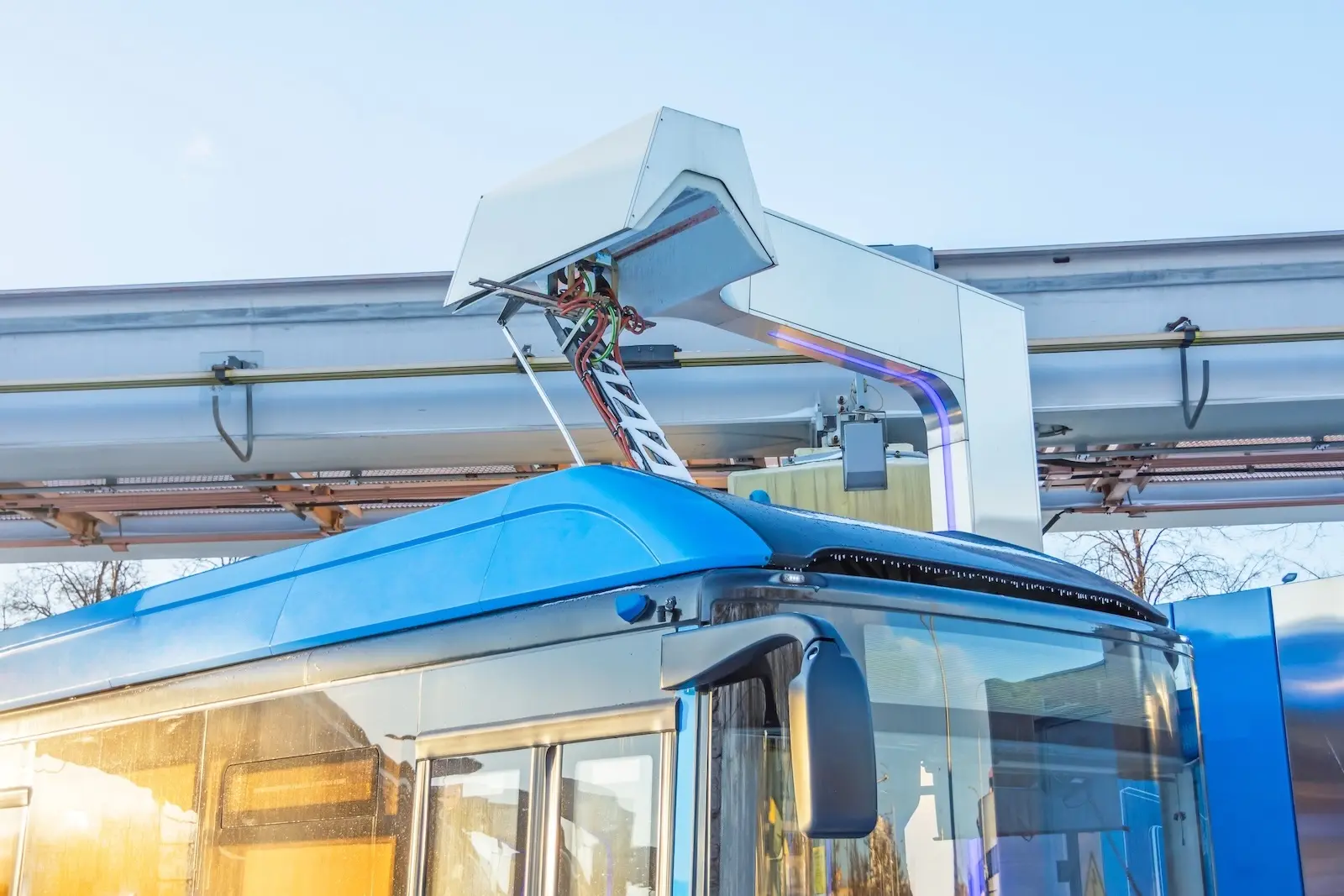 Blue electric bus connected to a white overhead charging station with cables and framework.