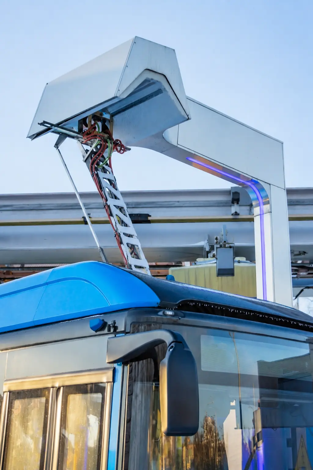 Blue electric bus connected to a white overhead charging station with cables and framework.