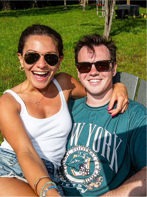 Smiling couple wearing sunglasses sitting outdoors on a sunny day with green grass in the background.