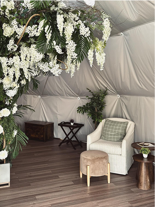 Cozy corner inside a tent with a white cushioned chair, green pillow, small brown ottoman, side tables, potted plants, and hanging white flowers.