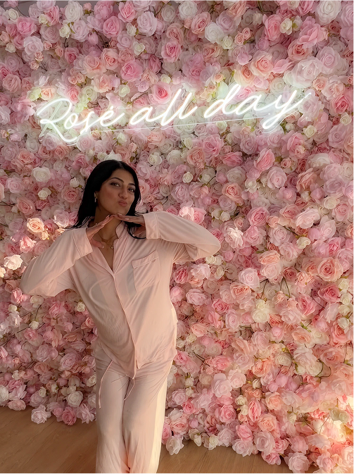 A woman in pink pajamas posing with hands under her chin in front of a wall covered with pink and white roses and a neon sign reading 'Rose all day'.