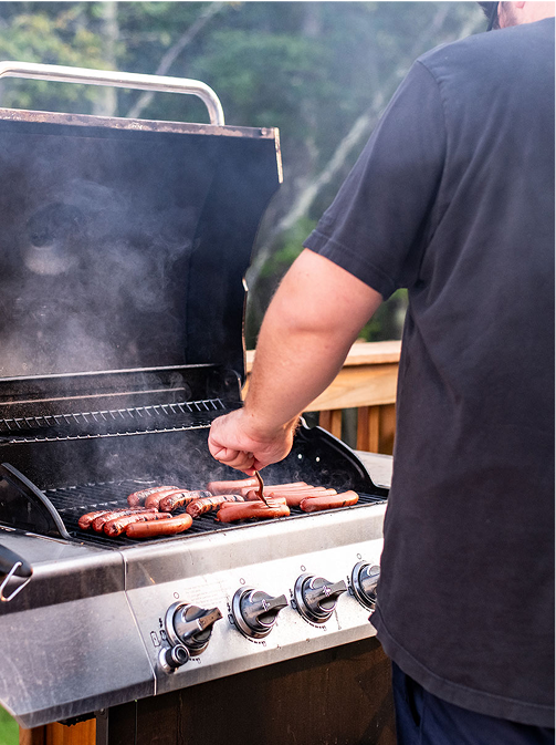 Person grilling multiple hot dogs on a gas barbecue grill outdoors with smoke rising.