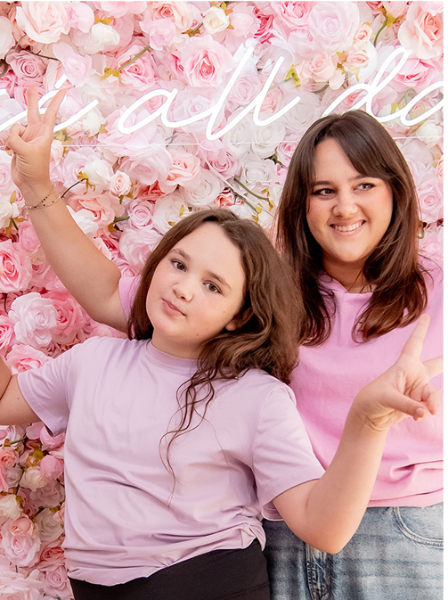 Two smiling females wearing light purple shirts making peace signs in front of a pink rose flower wall.