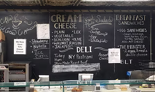 A chalkboard menu behind a deli counter listing cream cheese options, breakfast sandwiches, and quick bites, representing the go-to local stop for freshly baked bagels and morning favorites.