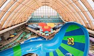 A panoramic view of New York's largest indoor waterpark under a vaulted wooden ceiling, featuring a large blue and green water slide, pools, and play areas, highlighting the top family fun and attractions.