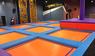 A large indoor room filled with interconnected trampolines, a climbing wall, and a sign for Laser Tag, representing the center for active indoor fun, including arcade games and family activities.