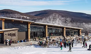 The modern base lodge and busy outdoor patio of the premier Catskills ski resort, with snow-covered mountains in the background, representing the four-season mountain adventure, ski slopes, and scenic hiking.