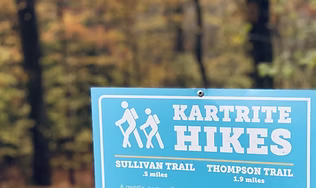 A close-up of a blue trail sign for 'Kartrite Hikes,' listing several named paths, representing the five peaceful nature trails available for a relaxed walk with varying lengths.