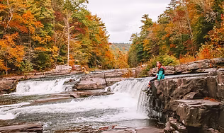 A scenic view of wide, cascading waterfalls flowing over dark, rocky terrain with vibrant autumn foliage in the background, highlighting the rewarding adventure on the 8.4-mile loop trail.